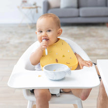 Load image into Gallery viewer, A baby in a high chair wearing a Bumkins Silicone Bib: Camp Gear eagerly tackles mealtime, enjoying food from a gray bowl with a spoon.