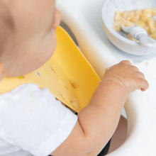 Load image into Gallery viewer, Close-up of a baby wearing a Bumkins Silicone Bib: Camp Gear, with mashed food in a bowl with a spoon nearby.