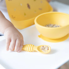 Load image into Gallery viewer, Close-up of a child wearing Bumkins Silicone Bib: Camp Gear, gripping a yellow spoon with food beside a matching bowl on a high chair tray.