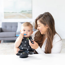 Load image into Gallery viewer, A woman smiles at a baby wearing a Bumkins Silicone Bib: Mickey Mouse Faces chewing a blue spoon in a bright, art-filled room.