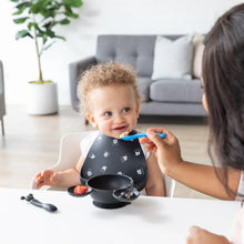 Load image into Gallery viewer, A baby with curly hair wears a Bumkins Silicone Bib: Mickey Mouse Faces, being fed by an adult at a table with a sofa and plant behind.