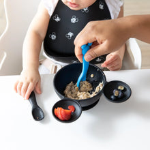 Load image into Gallery viewer, A baby wears a Bumkins Silicone Bib: Mickey Mouse Faces, while an adult hand used a blue spoon to scoop oatmeal from a black bowl.