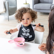 Load image into Gallery viewer, A curly haired child holds Bumkins Silicone Dipping Spoons: Minnie Mouse (Black and Pink) over a pink Minnie Mouse-shaped bowl of food.
