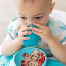 Load image into Gallery viewer, Close-up of a baby in a bib drinking from a Bumkins Silicone Starter Cup: Blue by a bowl of oatmeal.