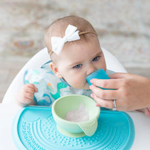 Load image into Gallery viewer, A baby in a bib and bow sits in a high chair learning to drink from a Bumkins Silicone Starter Cup: Blue, guided by an adult hand.