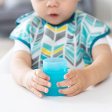 Load image into Gallery viewer, A baby in a patterned bib holds a Bumkins Silicone Starter Cup: Blue, seated happily at a white table.