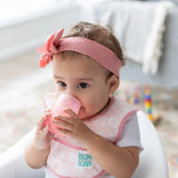 A baby wearing a pink headband and bib in a high chair drinks from a Bumkins Silicone Starter Cup: Pink.
