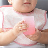 Close-up of a baby in a pink patterned bib holding a Bumkins Silicone Starter Cup: Pink.
