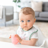 A baby with blue eyes in a floral bib sits in a high chair, holding a Bumkins Silicone Starter Cup: Pink.