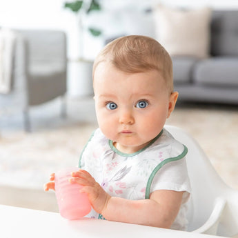 A baby with blue eyes in a floral bib sits in a high chair, holding a Bumkins Silicone Starter Cup: Pink.