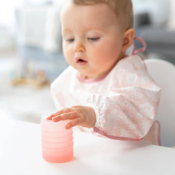 A baby in a pink patterned bib reaches for a Bumkins Silicone Starter Cup: Pink on a white table.