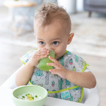 Load image into Gallery viewer, A baby drinks from a Bumkins Silicone Starter Cup: Sage, wearing a cactus bib, with a bowl of food on a high chair tray.