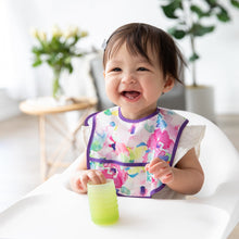 Load image into Gallery viewer, A smiling girl in a high chair wearing a floral bib holds a Bumkins Silicone Starter Cup: Sage in a bright room with plants.