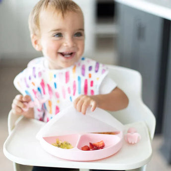 A toddler with a colorful bib in a high chair lifts a Bumkins Silicone Stretch Lid for Grip Dish 3 Section, Grip Plate from a pink plate.