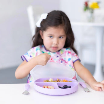 A girl with a floral bib holds a Bumkins Silicone Stretch Lid for Grip Dish 3 Section, Grip Plate over a purple plate with food on a table.