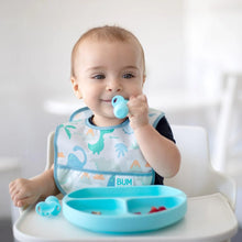 Load image into Gallery viewer, A baby in a high chair wears a Bumkins Starter Bib 2 Pack: Dinosaurs & Blue Tropic, eating from a blue divided plate with blue baby utensils.