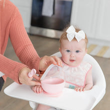 Load image into Gallery viewer, A baby in high chair with white bow wears a Bumkins Starter Bib 2 Pack: Floral & Lace as an adult places a pink bowl on the tray.