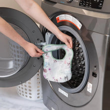 Load image into Gallery viewer, A person holds a Bumkins Starter Bib 2 Pack: Floral & Lace in front of an open front-loading washing machine.