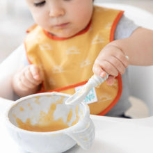 Load image into Gallery viewer, A baby wearing a yellow Bumkins Starter Bib 2 Pack: Sunshine and Grounded in a high chair with a spoon, eating from a bowl of mushy food.
