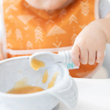 Load image into Gallery viewer, Close-up of a child in an orange Bumkins Starter Bib 2 Pack: Sunshine and Grounded, holding a spoon of baby food over a bowl.