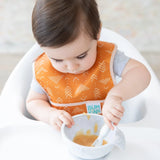 A toddler in a high chair eats from a gray bowl with a spoon while wearing an orange Bumkins Starter Bib 2 Pack: Sunshine and Grounded.