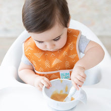 Load image into Gallery viewer, A toddler in a high chair eats from a gray bowl with a spoon while wearing an orange Bumkins Starter Bib 2 Pack: Sunshine and Grounded.