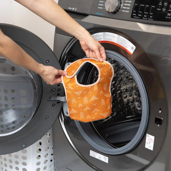 A person holds an orange Bumkins Starter Bib 2 Pack: Sunshine and Grounded in front of an open front-loading washing machine.