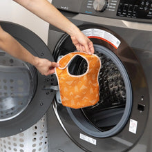 Load image into Gallery viewer, A person holds an orange Bumkins Starter Bib 2 Pack: Sunshine and Grounded in front of an open front-loading washing machine.