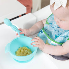 Load image into Gallery viewer, A baby in a Bumkins Starter Bib 2 Pack: Ocean Life & Whale Tail holds a spoon over a bowl of mashed food on a high chair tray.