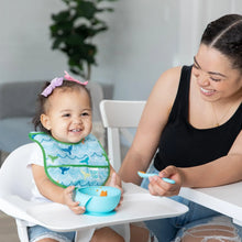 Load image into Gallery viewer, A smiling woman feeds a happy toddler in a high chair wearing a Bumkins Starter Bib 2 Pack: Ocean Life & Whale Tail from a blue bowl.