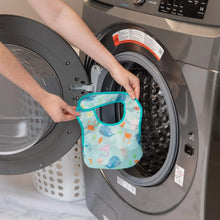 Load image into Gallery viewer, A person holds a Bumkins Starter Bib 2 Pack: Ocean Life & Whale Tail in front of an open front-loading washing machine.