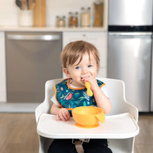 Load image into Gallery viewer, A toddler in a high chair, wearing a Bumkins Starter Bib 2 Pack: Jungle & Animal Prints, chews a spoon with an orange bowl nearby.