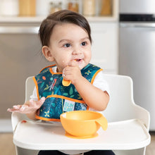 Load image into Gallery viewer, A baby in a Bumkins Starter Bib 2 Pack: Jungle & Animal Prints sits in a high chair, eating from an orange bowl with a spoon.