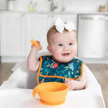 Load image into Gallery viewer, A baby sits in a high chair, wearing a Bumkins Starter Bib 2 Pack: Jungle & Animal Prints and holding an orange spoon near a matching bowl.