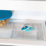 Close-up of a glass refrigerator shelf with a Bumkins Silicone Teething Charms: Blue beside a covered bowl of food.