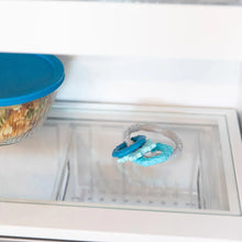 Load image into Gallery viewer, Close-up of a glass refrigerator shelf with a Bumkins Silicone Teething Charms: Blue beside a covered bowl of food.