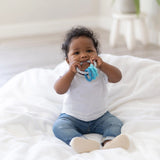 A baby, wearing a white shirt and jeans, chews on a Bumkins Silicone Teething Charms: Blue while sitting on a white blanket.