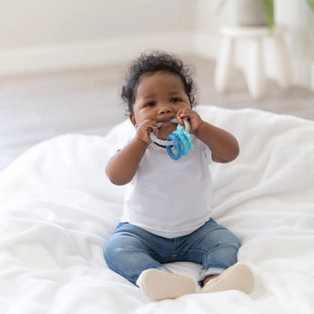 A baby, wearing a white shirt and jeans, chews on a Bumkins Silicone Teething Charms: Blue while sitting on a white blanket.