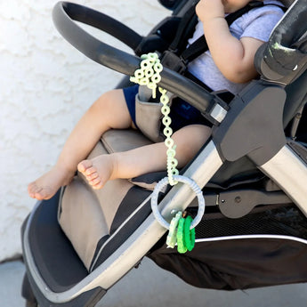 A baby sits in a stroller as a Bumkins Silicone Teething Charms: Green hangs from a green silicone tether attached to a handle.