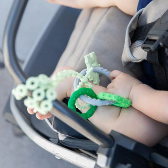 Close-up of a baby holding a Bumkins Silicone Teething Charms: Green while seated in a stroller.