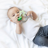 A baby lying on a white blanket, wearing a white shirt and gray pants and chewing a Bumkins Silicone Teething Charms: Green.