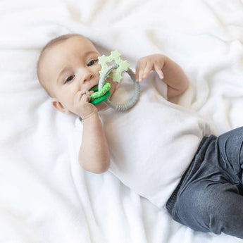 A baby lying on a white blanket, wearing a white shirt and gray pants and chewing a Bumkins Silicone Teething Charms: Green.