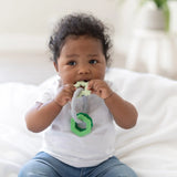 A baby in a white shirt and jeans sits on a white blanket, chewing on a Bumkins Silicone Teething Charms: Green.
