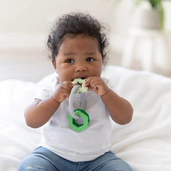 A baby in a white shirt and jeans sits on a white blanket, chewing on a Bumkins Silicone Teething Charms: Green.