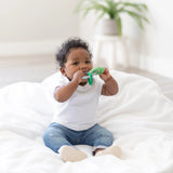 A curly-haired toddler sits on a white blanket, wearing a white shirt and jeans, while chewing a Bumkins Silicone Teething Charms: Green.