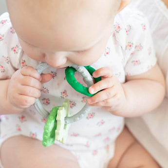 Close-up of a baby in a floral onesie chewing on a Bumkins Silicone Teething Charms: Green.