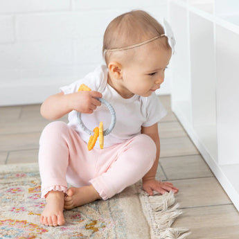 A baby in a white bow headband sits on a rug, holding a Bumkins Silicone Teething Charms: Orange by white shelves.