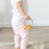 A toddler in a white shirt and pink pants holds Bumkins Silicone Teething Charms: Orange, standing on a rug in a bright room.