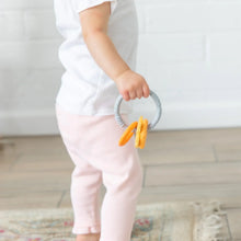Load image into Gallery viewer, A toddler in a white shirt and pink pants holds Bumkins Silicone Teething Charms: Orange, standing on a rug in a bright room.