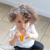 A toddler with curly hair in a white shirt and black pants chews on a Bumkins Silicone Teething Charms: Orange while seated on a wood floor.